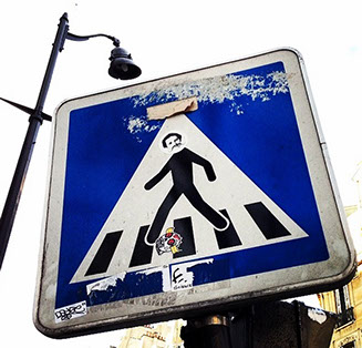 Blue, black, and white crosswalk sign in Paris, France. Has peeled off stickers and graffiti on top and a large street light behind it.